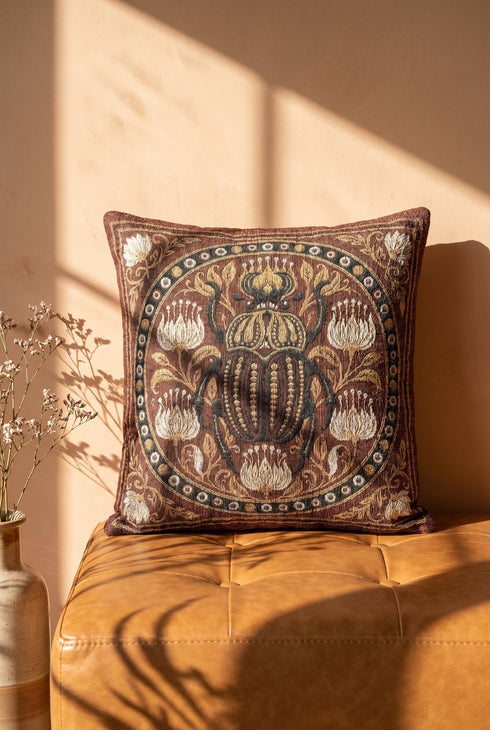 Square 17 by 18 inch jacquard woven throw pillow, woven identically on both faces and closed with a clean finished edge. A large detailed scarab beetle in black, gold, and dark brown sits at the center inside a dotted circular border, surrounded by symmetrical Art Nouveau vines and cream lotus flowers on a rust red ground. Small white floral motifs sit in each corner. The design is woven flat into the cotton blend cloth, with visible thread texture and a soft sheen in the ground.