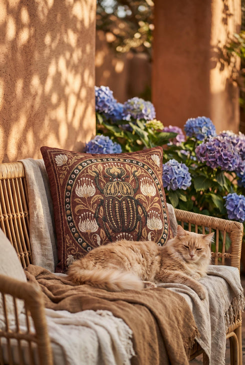 Square 17 by 18 inch jacquard woven throw pillow, woven identically on both faces and closed with a clean finished edge. A large detailed scarab beetle in black, gold, and dark brown sits at the center inside a dotted circular border, surrounded by symmetrical Art Nouveau vines and cream lotus flowers on a rust red ground. Small white floral motifs sit in each corner. The design is woven flat into the cotton blend cloth, with visible thread texture and a soft sheen in the ground.