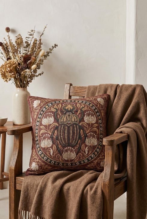 Square 17 by 18 inch jacquard woven throw pillow, woven identically on both faces and closed with a clean finished edge. A large detailed scarab beetle in black, gold, and dark brown sits at the center inside a dotted circular border, surrounded by symmetrical Art Nouveau vines and cream lotus flowers on a rust red ground. Small white floral motifs sit in each corner. The design is woven flat into the cotton blend cloth, with visible thread texture and a soft sheen in the ground.