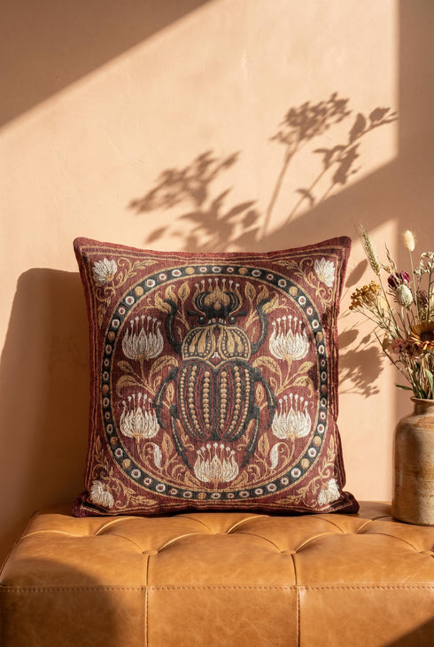 Square 17 by 18 inch jacquard woven throw pillow, woven identically on both faces and closed with a clean finished edge. A large detailed scarab beetle in black, gold, and dark brown sits at the center inside a dotted circular border, surrounded by symmetrical Art Nouveau vines and cream lotus flowers on a rust red ground. Small white floral motifs sit in each corner. The design is woven flat into the cotton blend cloth, with visible thread texture and a soft sheen in the ground.