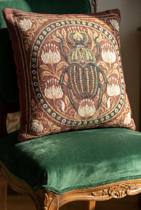 Square 17 by 18 inch jacquard woven throw pillow, woven identically on both faces and closed with a clean finished edge. A large detailed scarab beetle in black, gold, and dark brown sits at the center inside a dotted circular border, surrounded by symmetrical Art Nouveau vines and cream lotus flowers on a rust red ground. Small white floral motifs sit in each corner. The design is woven flat into the cotton blend cloth, with visible thread texture and a soft sheen in the ground.