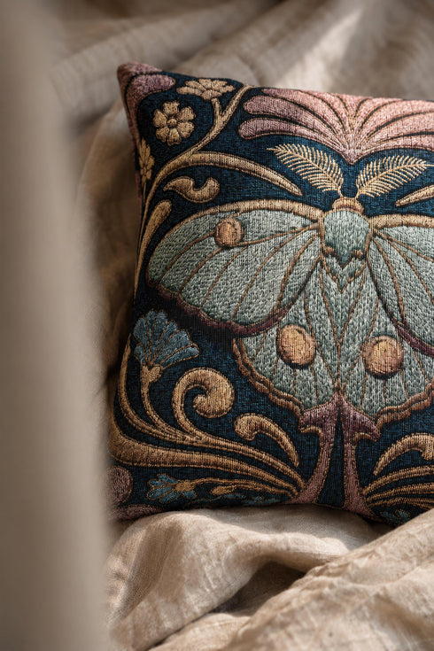 Square 17 by 18 inch jacquard woven throw pillow, woven identically on both faces and closed with a clean finished edge. A symmetrical Art Nouveau luna moth in pale sage green spreads its wings at the center of a deep teal ground. Gold scrollwork, dusty mauve palmette fans, French blue blossoms, and small cream flowers frame the moth. The design is woven flat into the cotton blend cloth, with visible thread texture across the motif and a soft sheen in the ground weave.