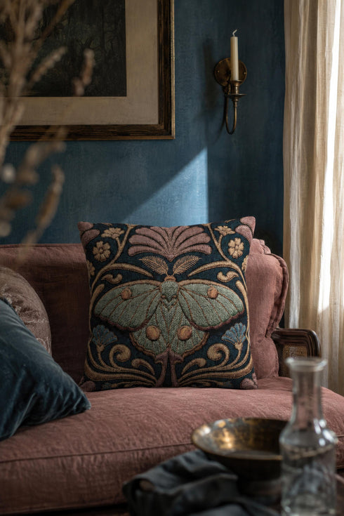 Square 17 by 18 inch jacquard woven throw pillow, woven identically on both faces and closed with a clean finished edge. A symmetrical Art Nouveau luna moth in pale sage green spreads its wings at the center of a deep teal ground. Gold scrollwork, dusty mauve palmette fans, French blue blossoms, and small cream flowers frame the moth. The design is woven flat into the cotton blend cloth, with visible thread texture across the motif and a soft sheen in the ground weave.