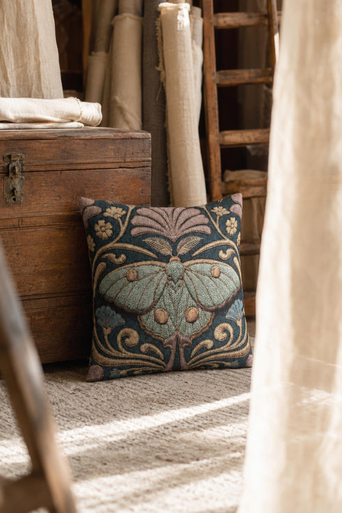 Square 17 by 18 inch jacquard woven throw pillow, woven identically on both faces and closed with a clean finished edge. A symmetrical Art Nouveau luna moth in pale sage green spreads its wings at the center of a deep teal ground. Gold scrollwork, dusty mauve palmette fans, French blue blossoms, and small cream flowers frame the moth. The design is woven flat into the cotton blend cloth, with visible thread texture across the motif and a soft sheen in the ground weave.