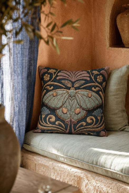 Square 17 by 18 inch jacquard woven throw pillow, woven identically on both faces and closed with a clean finished edge. A symmetrical Art Nouveau luna moth in pale sage green spreads its wings at the center of a deep teal ground. Gold scrollwork, dusty mauve palmette fans, French blue blossoms, and small cream flowers frame the moth. The design is woven flat into the cotton blend cloth, with visible thread texture across the motif and a soft sheen in the ground weave.