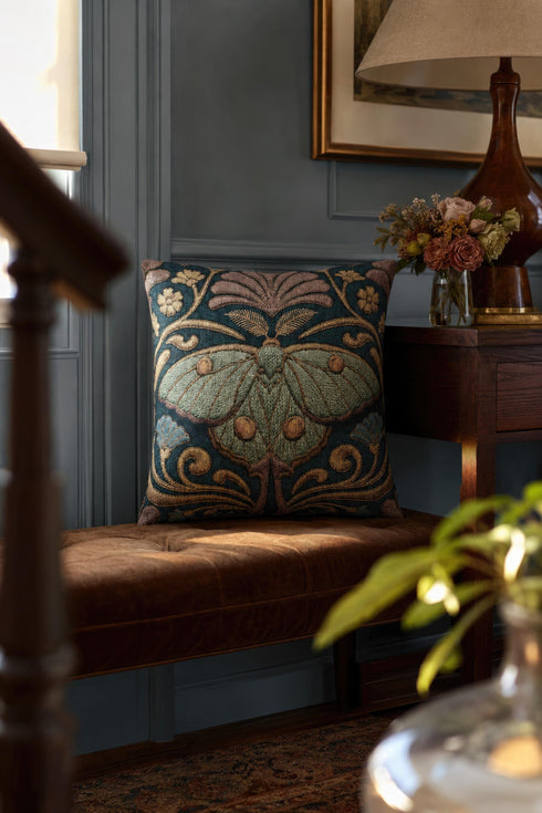 Square 17 by 18 inch jacquard woven throw pillow, woven identically on both faces and closed with a clean finished edge. A symmetrical Art Nouveau luna moth in pale sage green spreads its wings at the center of a deep teal ground. Gold scrollwork, dusty mauve palmette fans, French blue blossoms, and small cream flowers frame the moth. The design is woven flat into the cotton blend cloth, with visible thread texture across the motif and a soft sheen in the ground weave.