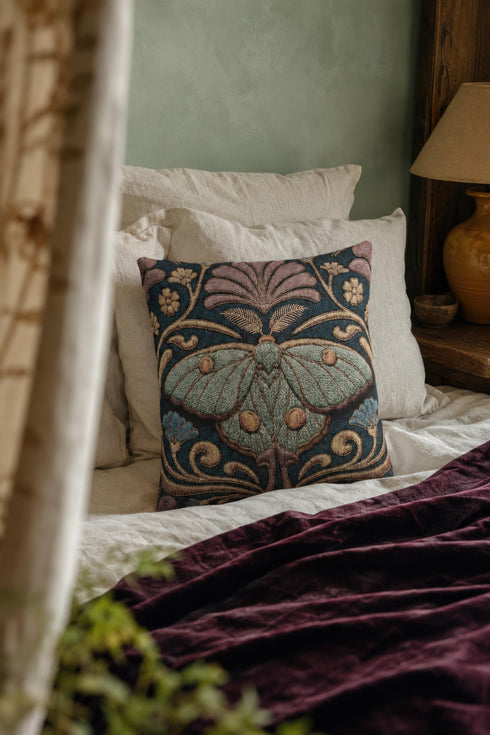 Square 17 by 18 inch jacquard woven throw pillow, woven identically on both faces and closed with a clean finished edge. A symmetrical Art Nouveau luna moth in pale sage green spreads its wings at the center of a deep teal ground. Gold scrollwork, dusty mauve palmette fans, French blue blossoms, and small cream flowers frame the moth. The design is woven flat into the cotton blend cloth, with visible thread texture across the motif and a soft sheen in the ground weave.