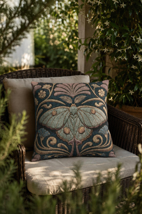 Square 17 by 18 inch jacquard woven throw pillow, woven identically on both faces and closed with a clean finished edge. A symmetrical Art Nouveau luna moth in pale sage green spreads its wings at the center of a deep teal ground. Gold scrollwork, dusty mauve palmette fans, French blue blossoms, and small cream flowers frame the moth. The design is woven flat into the cotton blend cloth, with visible thread texture across the motif and a soft sheen in the ground weave.