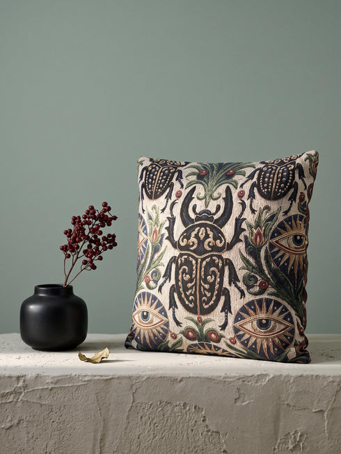 Square 17 by 18 inch jacquard woven throw pillow, woven identically on both faces and closed with a clean finished edge. On a natural cream ecru ground, a horned scarab beetle with gold filigree scrollwork on its shell sits at the center, flanked by two evil eye medallions with sapphire blue centers in sunburst halos. Two smaller scarabs, sage green acanthus vines, and crimson berries frame the edges. The design is woven flat into the cotton blend cloth with visible thread texture.
