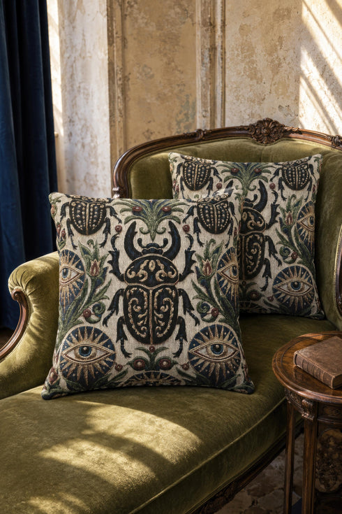Square 17 by 18 inch jacquard woven throw pillow, woven identically on both faces and closed with a clean finished edge. On a natural cream ecru ground, a horned scarab beetle with gold filigree scrollwork on its shell sits at the center, flanked by two evil eye medallions with sapphire blue centers in sunburst halos. Two smaller scarabs, sage green acanthus vines, and crimson berries frame the edges. The design is woven flat into the cotton blend cloth with visible thread texture.