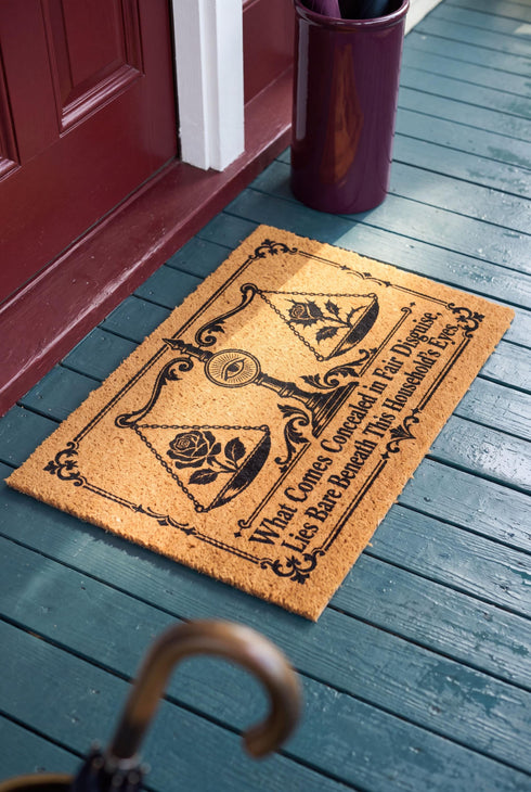 Rectangular 24 by 16 inch tufted natural coconut fiber coir doormat with squared corners and a black vinyl backing. The black artwork is printed into the fibers: an ornate antique balance scale with two suspended pans, each holding a blooming rose stem. A radiant eye in an oval sunburst crests the center column. Ornate scrollwork frames the lower inscription, which reads WHAT COMES CONCEALED IN FAIR DISGUISE, LIES BARE BENEATH THIS HOUSEHOLD'S EYES.
