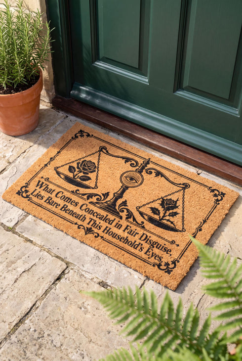 Rectangular 24 by 16 inch tufted natural coconut fiber coir doormat with squared corners and a black vinyl backing. The black artwork is printed into the fibers: an ornate antique balance scale with two suspended pans, each holding a blooming rose stem. A radiant eye in an oval sunburst crests the center column. Ornate scrollwork frames the lower inscription, which reads WHAT COMES CONCEALED IN FAIR DISGUISE, LIES BARE BENEATH THIS HOUSEHOLD'S EYES.