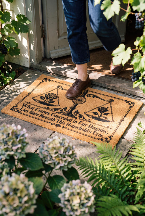 Rectangular 24 by 16 inch tufted natural coconut fiber coir doormat with squared corners and a black vinyl backing. The black artwork is printed into the fibers: an ornate antique balance scale with two suspended pans, each holding a blooming rose stem. A radiant eye in an oval sunburst crests the center column. Ornate scrollwork frames the lower inscription, which reads WHAT COMES CONCEALED IN FAIR DISGUISE, LIES BARE BENEATH THIS HOUSEHOLD'S EYES.