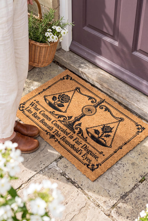Rectangular 24 by 16 inch tufted natural coconut fiber coir doormat with squared corners and a black vinyl backing. The black artwork is printed into the fibers: an ornate antique balance scale with two suspended pans, each holding a blooming rose stem. A radiant eye in an oval sunburst crests the center column. Ornate scrollwork frames the lower inscription, which reads WHAT COMES CONCEALED IN FAIR DISGUISE, LIES BARE BENEATH THIS HOUSEHOLD'S EYES.