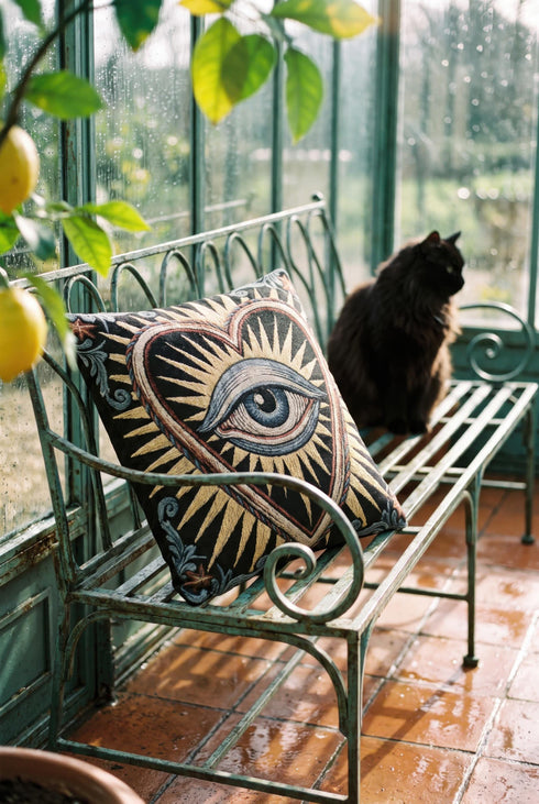Square 17 by 18 inch jacquard woven throw pillow, woven identically on both faces and closed with a clean finished edge. On a solid black ground, a large blue evil eye sits inside a golden sunburst heart. Gold, pale blue, cream, and rust threads form the design, with dusty blue acanthus leaves curling around the heart and small rust stars in each corner. The design is woven flat into the cotton blend cloth, with visible thread texture and a heavy tapestry hand.