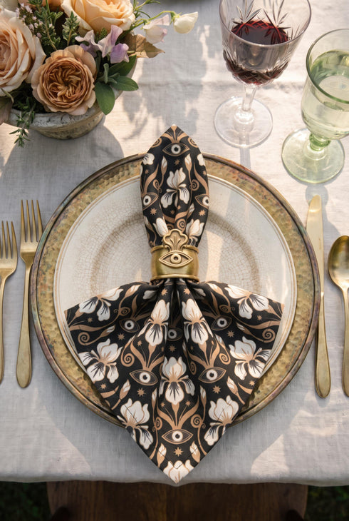 Set of four square cloth dinner napkins, each 20 by 20 inches, in polyester broadcloth with hemmed edges on all four sides. The top face is printed with a dense Art Nouveau pattern on a black ground: cream iris blossoms, curving gold toned vines, small starbursts, and evil eye talismans arranged in a damask like layout, with no lettering. The underside is white and shows the design faintly through the smooth, densely woven fabric.