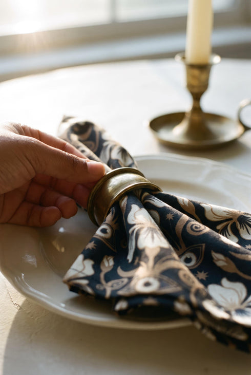 Set of four square cloth dinner napkins, each 20 by 20 inches, in polyester broadcloth with hemmed edges on all four sides. The top face is printed with a dense Art Nouveau pattern on a black ground: cream iris blossoms, curving gold toned vines, small starbursts, and evil eye talismans arranged in a damask like layout, with no lettering. The underside is white and shows the design faintly through the smooth, densely woven fabric.