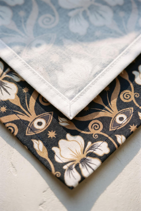 Set of four square cloth dinner napkins, each 20 by 20 inches, in polyester broadcloth with hemmed edges on all four sides. The top face is printed with a dense Art Nouveau pattern on a black ground: cream iris blossoms, curving gold toned vines, small starbursts, and evil eye talismans arranged in a damask like layout, with no lettering. The underside is white and shows the design faintly through the smooth, densely woven fabric.