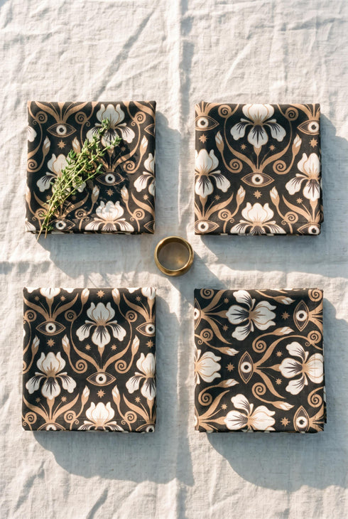 Set of four square cloth dinner napkins, each 20 by 20 inches, in polyester broadcloth with hemmed edges on all four sides. The top face is printed with a dense Art Nouveau pattern on a black ground: cream iris blossoms, curving gold toned vines, small starbursts, and evil eye talismans arranged in a damask like layout, with no lettering. The underside is white and shows the design faintly through the smooth, densely woven fabric.