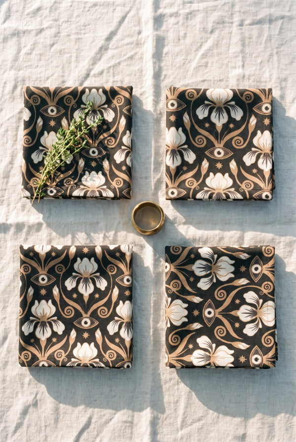 Set of four square cloth dinner napkins, each 20 by 20 inches, in polyester broadcloth with hemmed edges on all four sides. The top face is printed with a dense Art Nouveau pattern on a black ground: cream iris blossoms, curving gold toned vines, small starbursts, and evil eye talismans arranged in a damask like layout, with no lettering. The underside is white and shows the design faintly through the smooth, densely woven fabric.