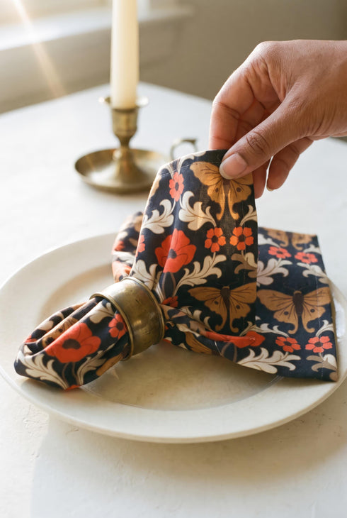 Set of four square cloth dinner napkins, each 20 by 20 inches, in polyester broadcloth with hemmed edges. The top face is printed with a mirrored damask style pattern of golden luna moths, burnt orange poppies, small coral blossoms, and ivory acanthus scrollwork on a solid black ground. The underside is white and shows a faint, softened echo of the printed pattern through the lightweight, smooth, densely woven fabric.
