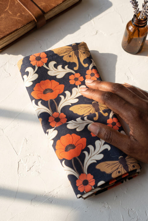 Set of four square cloth dinner napkins, each 20 by 20 inches, in polyester broadcloth with hemmed edges. The top face is printed with a mirrored damask style pattern of golden luna moths, burnt orange poppies, small coral blossoms, and ivory acanthus scrollwork on a solid black ground. The underside is white and shows a faint, softened echo of the printed pattern through the lightweight, smooth, densely woven fabric.