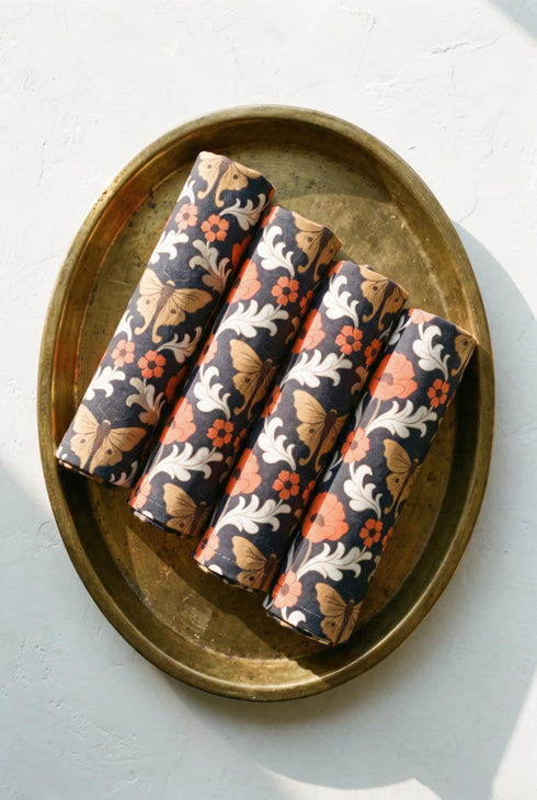 Set of four square cloth dinner napkins, each 20 by 20 inches, in polyester broadcloth with hemmed edges. The top face is printed with a mirrored damask style pattern of golden luna moths, burnt orange poppies, small coral blossoms, and ivory acanthus scrollwork on a solid black ground. The underside is white and shows a faint, softened echo of the printed pattern through the lightweight, smooth, densely woven fabric.