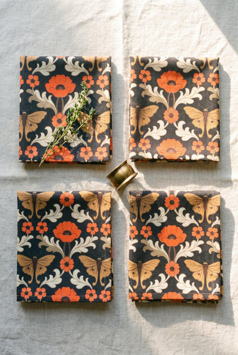 Set of four square cloth dinner napkins, each 20 by 20 inches, in polyester broadcloth with hemmed edges. The top face is printed with a mirrored damask style pattern of golden luna moths, burnt orange poppies, small coral blossoms, and ivory acanthus scrollwork on a solid black ground. The underside is white and shows a faint, softened echo of the printed pattern through the lightweight, smooth, densely woven fabric.