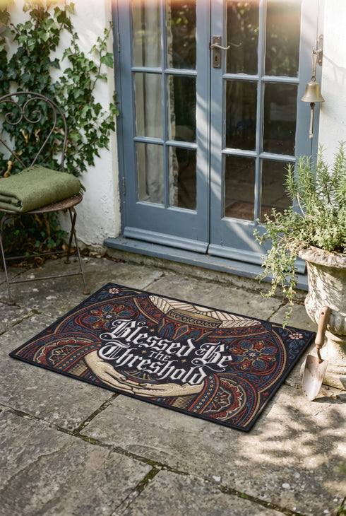 Rectangular navy doormat with a low loop polyester pile and black border trim, printed flat to imitate hand embroidery and crewelwork. Two pale hands offer a golden chalice at the center, with white rays streaming from the cup. Ornate white lettering across the design reads BLESSED BE THE THRESHOLD. Rust red and antique gold Arts and Crafts botanical arches frame the scene, curving over and around the chalice against the deep navy ground.