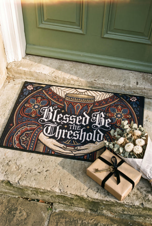 Rectangular navy doormat with a low loop polyester pile and black border trim, printed flat to imitate hand embroidery and crewelwork. Two pale hands offer a golden chalice at the center, with white rays streaming from the cup. Ornate white lettering across the design reads BLESSED BE THE THRESHOLD. Rust red and antique gold Arts and Crafts botanical arches frame the scene, curving over and around the chalice against the deep navy ground.