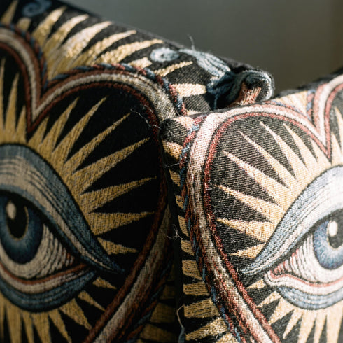 Square 17 by 18 inch jacquard woven throw pillow, woven identically on both faces and closed with a clean finished edge. On a solid black ground, a large blue evil eye sits inside a golden sunburst heart. Gold, pale blue, cream, and rust threads form the design, with dusty blue acanthus leaves curling around the heart and small rust stars in each corner. The design is woven flat into the cotton blend cloth, with visible thread texture and a heavy tapestry hand.