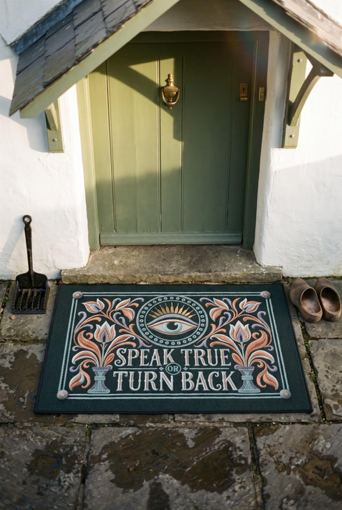 Rectangular doormat with a deep evergreen ground, low loop polyester pile, and black edge trim, printed flat edge to edge to imitate raised embroidery. A central eye with a lavender lid, coral outline, and seafoam iris sits inside a beaded circle under gold sunburst rays. Coral, lavender, and cream folk art tulips rise from seafoam urns on each side. Cream lettering with coral inlines reads SPEAK TRUE OR TURN BACK, framed by a seafoam rope border.