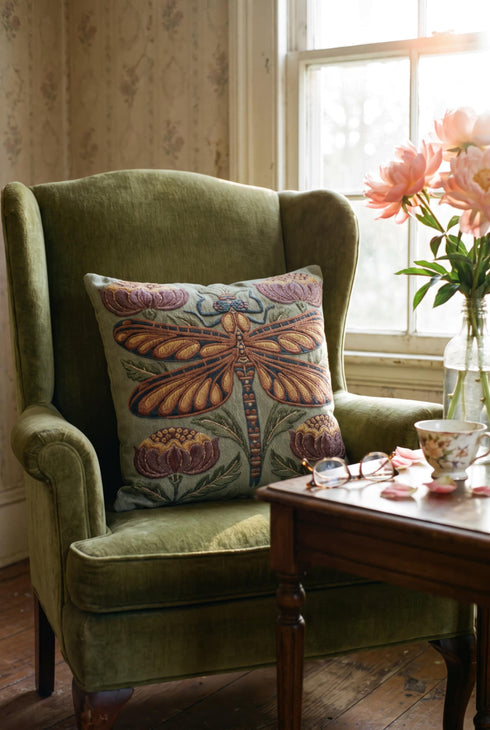 Square 17 by 18 inch jacquard woven throw pillow, woven identically on both faces and closed with a clean finished edge. A mirror symmetrical dragonfly fills the center of a sage green ground, its four long wings outlined in rust copper and filled with apricot and mustard, barred in slate blue along the leading edge. Plum peonies with ochre centers sit in each corner, and olive leaf sprays rise from the base. The ground has a soft sheen and the motif shows matte woven thread.