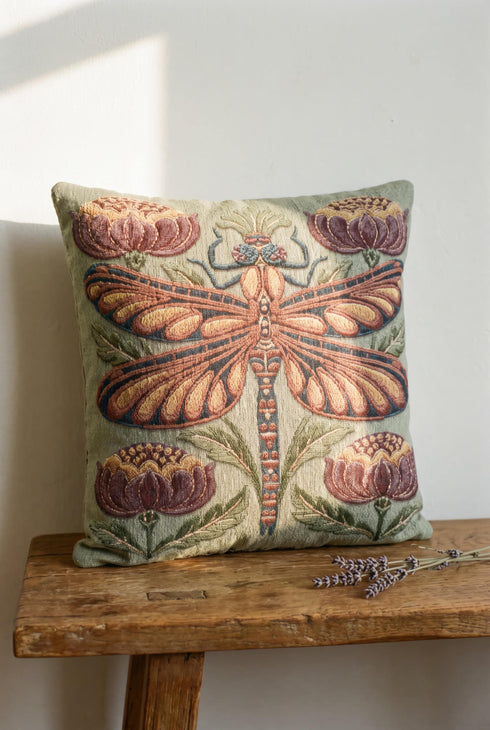 Square 17 by 18 inch jacquard woven throw pillow, woven identically on both faces and closed with a clean finished edge. A mirror symmetrical dragonfly fills the center of a sage green ground, its four long wings outlined in rust copper and filled with apricot and mustard, barred in slate blue along the leading edge. Plum peonies with ochre centers sit in each corner, and olive leaf sprays rise from the base. The ground has a soft sheen and the motif shows matte woven thread.