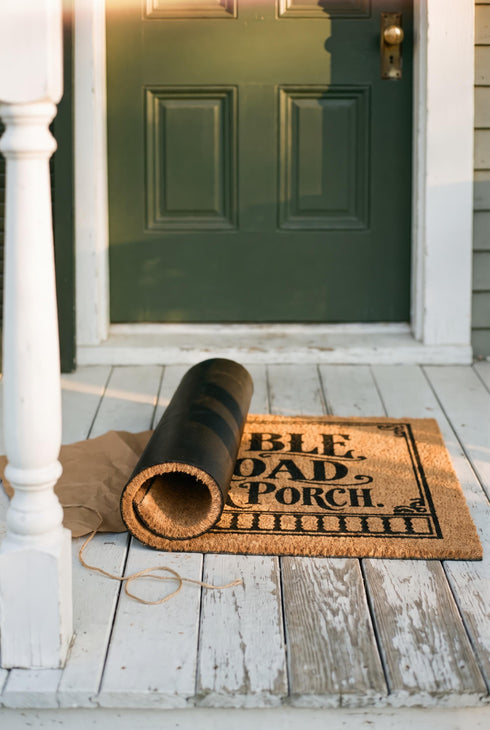 Rectangular natural tan coir doormat, 24 by 16 inches, with squared corners, a bristled tufted coconut fiber surface, and a black vinyl backing. The black artwork is printed into the fibers inside a double line frame. Ornate Victorian lettering reads MAY TROUBLE TAKE THE ROAD THAT NEVER FINDS OUR PORCH. Turned porch posts with scrolled corner brackets stand on each side, a row of balusters runs along the bottom, and a small winding road and magnolia blossom sit at the center.