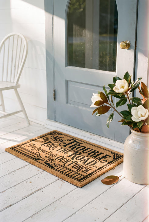 Rectangular natural tan coir doormat, 24 by 16 inches, with squared corners, a bristled tufted coconut fiber surface, and a black vinyl backing. The black artwork is printed into the fibers inside a double line frame. Ornate Victorian lettering reads MAY TROUBLE TAKE THE ROAD THAT NEVER FINDS OUR PORCH. Turned porch posts with scrolled corner brackets stand on each side, a row of balusters runs along the bottom, and a small winding road and magnolia blossom sit at the center.