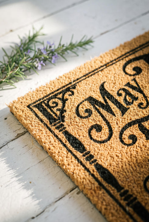 Rectangular natural tan coir doormat, 24 by 16 inches, with squared corners, a bristled tufted coconut fiber surface, and a black vinyl backing. The black artwork is printed into the fibers inside a double line frame. Ornate Victorian lettering reads MAY TROUBLE TAKE THE ROAD THAT NEVER FINDS OUR PORCH. Turned porch posts with scrolled corner brackets stand on each side, a row of balusters runs along the bottom, and a small winding road and magnolia blossom sit at the center.