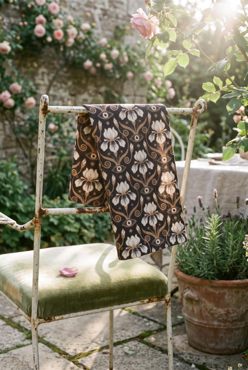 Set of four square cloth dinner napkins, each 20 by 20 inches, in polyester broadcloth with hemmed edges on all four sides. The top face is printed with a dense Art Nouveau pattern on a black ground: cream iris blossoms, curving gold toned vines, small starbursts, and evil eye talismans arranged in a damask like layout, with no lettering. The underside is white and shows the design faintly through the smooth, densely woven fabric.