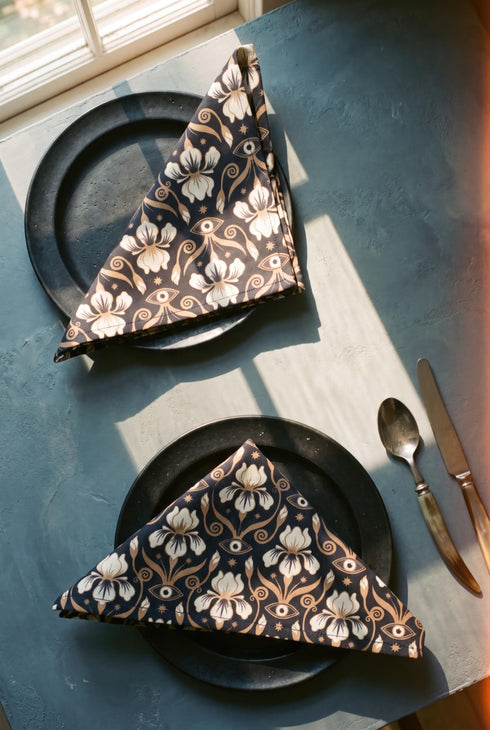 Set of four square cloth dinner napkins, each 20 by 20 inches, in polyester broadcloth with hemmed edges on all four sides. The top face is printed with a dense Art Nouveau pattern on a black ground: cream iris blossoms, curving gold toned vines, small starbursts, and evil eye talismans arranged in a damask like layout, with no lettering. The underside is white and shows the design faintly through the smooth, densely woven fabric.
