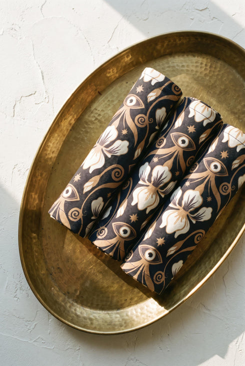 Set of four square cloth dinner napkins, each 20 by 20 inches, in polyester broadcloth with hemmed edges on all four sides. The top face is printed with a dense Art Nouveau pattern on a black ground: cream iris blossoms, curving gold toned vines, small starbursts, and evil eye talismans arranged in a damask like layout, with no lettering. The underside is white and shows the design faintly through the smooth, densely woven fabric.