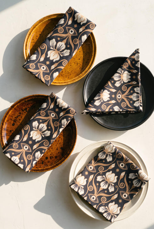 Set of four square cloth dinner napkins, each 20 by 20 inches, in polyester broadcloth with hemmed edges on all four sides. The top face is printed with a dense Art Nouveau pattern on a black ground: cream iris blossoms, curving gold toned vines, small starbursts, and evil eye talismans arranged in a damask like layout, with no lettering. The underside is white and shows the design faintly through the smooth, densely woven fabric.