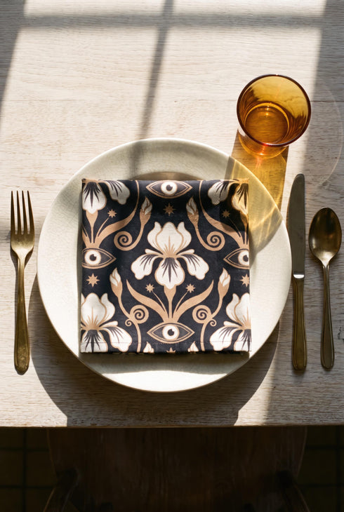 Set of four square cloth dinner napkins, each 20 by 20 inches, in polyester broadcloth with hemmed edges on all four sides. The top face is printed with a dense Art Nouveau pattern on a black ground: cream iris blossoms, curving gold toned vines, small starbursts, and evil eye talismans arranged in a damask like layout, with no lettering. The underside is white and shows the design faintly through the smooth, densely woven fabric.