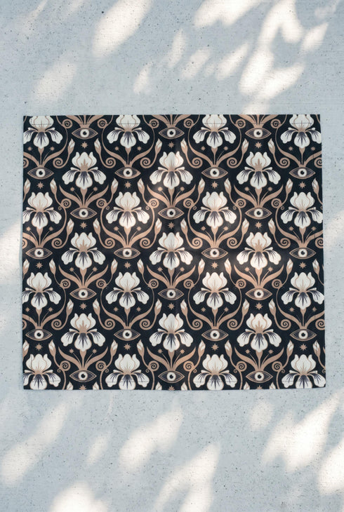 Set of four square cloth dinner napkins, each 20 by 20 inches, in polyester broadcloth with hemmed edges on all four sides. The top face is printed with a dense Art Nouveau pattern on a black ground: cream iris blossoms, curving gold toned vines, small starbursts, and evil eye talismans arranged in a damask like layout, with no lettering. The underside is white and shows the design faintly through the smooth, densely woven fabric.