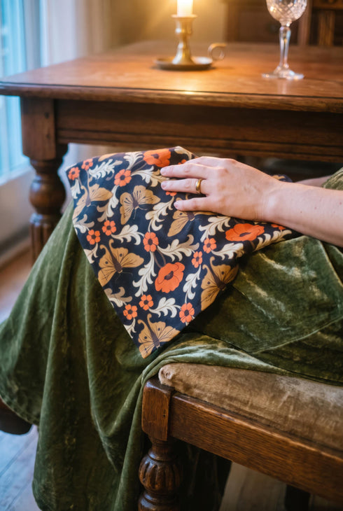 Set of four square cloth dinner napkins, each 20 by 20 inches, in polyester broadcloth with hemmed edges. The top face is printed with a mirrored damask style pattern of golden luna moths, burnt orange poppies, small coral blossoms, and ivory acanthus scrollwork on a solid black ground. The underside is white and shows a faint, softened echo of the printed pattern through the lightweight, smooth, densely woven fabric.