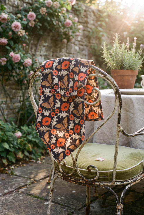 Set of four square cloth dinner napkins, each 20 by 20 inches, in polyester broadcloth with hemmed edges. The top face is printed with a mirrored damask style pattern of golden luna moths, burnt orange poppies, small coral blossoms, and ivory acanthus scrollwork on a solid black ground. The underside is white and shows a faint, softened echo of the printed pattern through the lightweight, smooth, densely woven fabric.