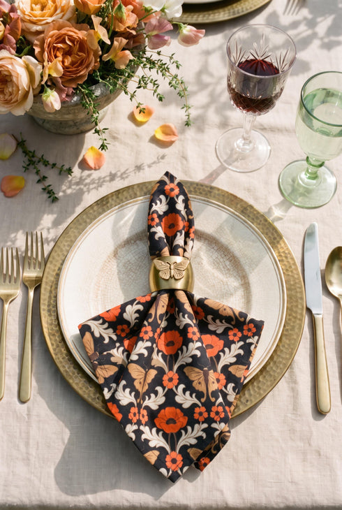 Set of four square cloth dinner napkins, each 20 by 20 inches, in polyester broadcloth with hemmed edges. The top face is printed with a mirrored damask style pattern of golden luna moths, burnt orange poppies, small coral blossoms, and ivory acanthus scrollwork on a solid black ground. The underside is white and shows a faint, softened echo of the printed pattern through the lightweight, smooth, densely woven fabric.