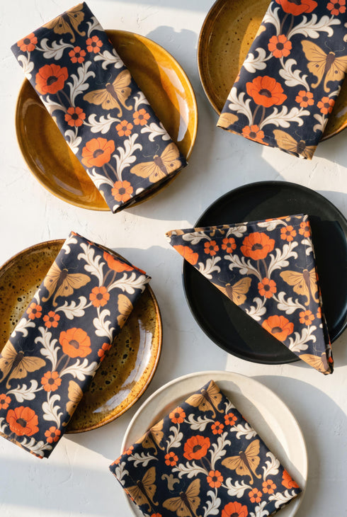 Set of four square cloth dinner napkins, each 20 by 20 inches, in polyester broadcloth with hemmed edges. The top face is printed with a mirrored damask style pattern of golden luna moths, burnt orange poppies, small coral blossoms, and ivory acanthus scrollwork on a solid black ground. The underside is white and shows a faint, softened echo of the printed pattern through the lightweight, smooth, densely woven fabric.