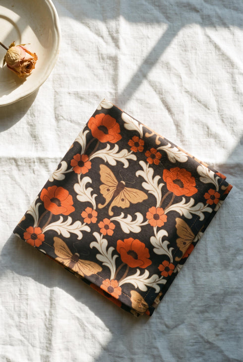 Set of four square cloth dinner napkins, each 20 by 20 inches, in polyester broadcloth with hemmed edges. The top face is printed with a mirrored damask style pattern of golden luna moths, burnt orange poppies, small coral blossoms, and ivory acanthus scrollwork on a solid black ground. The underside is white and shows a faint, softened echo of the printed pattern through the lightweight, smooth, densely woven fabric.