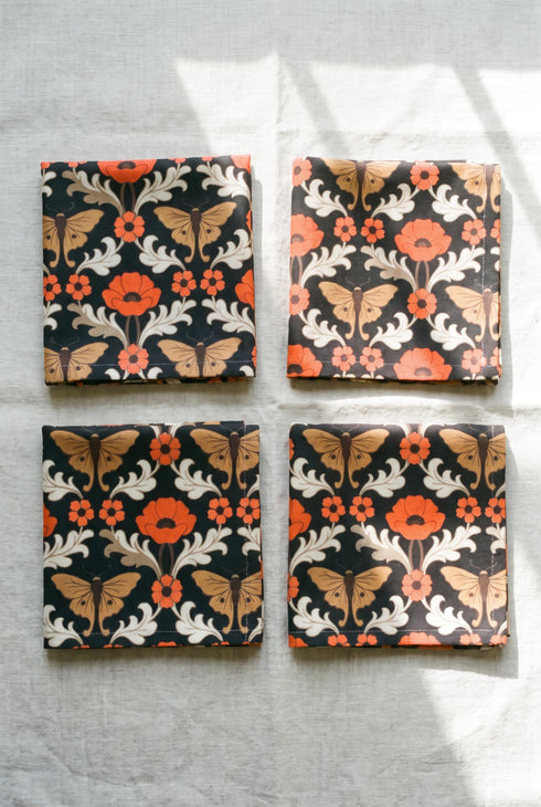 Set of four square cloth dinner napkins, each 20 by 20 inches, in polyester broadcloth with hemmed edges. The top face is printed with a mirrored damask style pattern of golden luna moths, burnt orange poppies, small coral blossoms, and ivory acanthus scrollwork on a solid black ground. The underside is white and shows a faint, softened echo of the printed pattern through the lightweight, smooth, densely woven fabric.