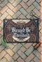 Rectangular navy doormat with a low loop polyester pile and black border trim, printed flat to imitate hand embroidery and crewelwork. Two pale hands offer a golden chalice at the center, with white rays streaming from the cup. Ornate white lettering across the design reads BLESSED BE THE THRESHOLD. Rust red and antique gold Arts and Crafts botanical arches frame the scene, curving over and around the chalice against the deep navy ground.